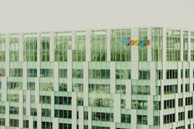 A modern office building with a glass facade reflecting the surroundings. The Google logo is prominently displayed on the exterior. The architecture features repetitive rectangular patterns with varying shades of green and white panels.