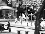 A black and white photo of an outdoor urban area featuring playground equipment. Several figures, mostly women and children, interact with the equipment. One woman is seen sitting on a bench in the foreground, while another interacts with children at a play apparatus. There is a parked scooter to the left and a blurred figure walking past. The area is shaded by large trees and the background shows buildings with various urban fixtures like air conditioning units.