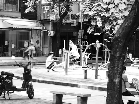 A black and white photo of an outdoor urban area featuring playground equipment. Several figures, mostly women and children, interact with the equipment. One woman is seen sitting on a bench in the foreground, while another interacts with children at a play apparatus. There is a parked scooter to the left and a blurred figure walking past. The area is shaded by large trees and the background shows buildings with various urban fixtures like air conditioning units.