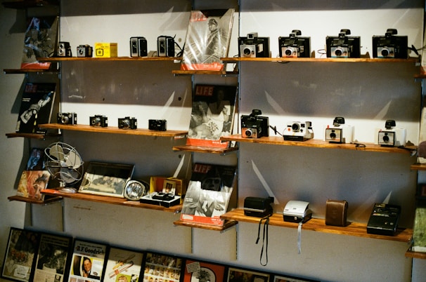 A collection of vintage cameras and magazines displayed on wooden shelves. The shelves are filled with various models of old cameras, alongside issues of LIFE magazine. Some small retro gadgets and books are also placed on the shelves.