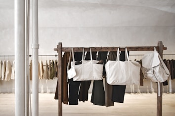 Several beige and neutral-colored bags are hanging from a wooden rack in a minimalist, modern space. Behind the rack, various neutral-toned jackets and pants are hanging against a light-colored wall. Two vertical white poles are positioned to the left.