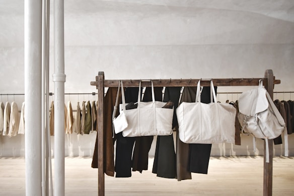 Several beige and neutral-colored bags are hanging from a wooden rack in a minimalist, modern space. Behind the rack, various neutral-toned jackets and pants are hanging against a light-colored wall. Two vertical white poles are positioned to the left.