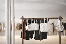 Several beige and neutral-colored bags are hanging from a wooden rack in a minimalist, modern space. Behind the rack, various neutral-toned jackets and pants are hanging against a light-colored wall. Two vertical white poles are positioned to the left.