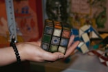 A hand holding an old, worn Rubik's Cube, with faded and peeling stickers. The background is cluttered with various colorful objects and patterns.
