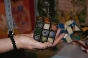 A hand holding an old, worn Rubik's Cube, with faded and peeling stickers. The background is cluttered with various colorful objects and patterns.