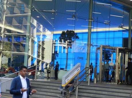 A glass-fronted modern office building with a bright blue interior lighting. The entrance features a revolving door and several people are visible, some entering or exiting. The facade reflects surrounding buildings and trees, creating an urban atmosphere. Steps lead up to the entrance, and a person in a suit is walking briskly in the foreground.