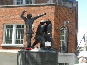A bronze statue depicting three soldiers in dynamic poses. Two figures appear to be supporting a third, highlighting a moment of camaraderie or assistance. The statue is set against a backdrop of a brick building with white-framed windows.