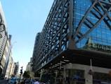 A modern, glass-fronted building with a grid-like structure dominates the foreground. The building features reflective blue-tinted windows and grey metal supports. To the left, an older style of architecture with intricate details lines the street. A clear blue sky provides a bright backdrop. A busy street scene includes people walking and the iconic London Underground sign near a station entrance.