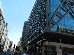 A modern, glass-fronted building with a grid-like structure dominates the foreground. The building features reflective blue-tinted windows and grey metal supports. To the left, an older style of architecture with intricate details lines the street. A clear blue sky provides a bright backdrop. A busy street scene includes people walking and the iconic London Underground sign near a station entrance.