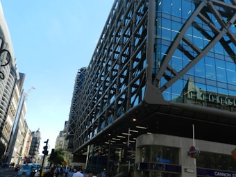 A modern, glass-fronted building with a grid-like structure dominates the foreground. The building features reflective blue-tinted windows and grey metal supports. To the left, an older style of architecture with intricate details lines the street. A clear blue sky provides a bright backdrop. A busy street scene includes people walking and the iconic London Underground sign near a station entrance.