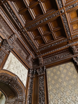 A detailed view of an ornately carved wooden ceiling with intricate patterns and designs. The woodwork includes floral and geometric motifs, covering the ceiling and archway. The walls feature elegant patterned wallpaper in a sophisticated setting.