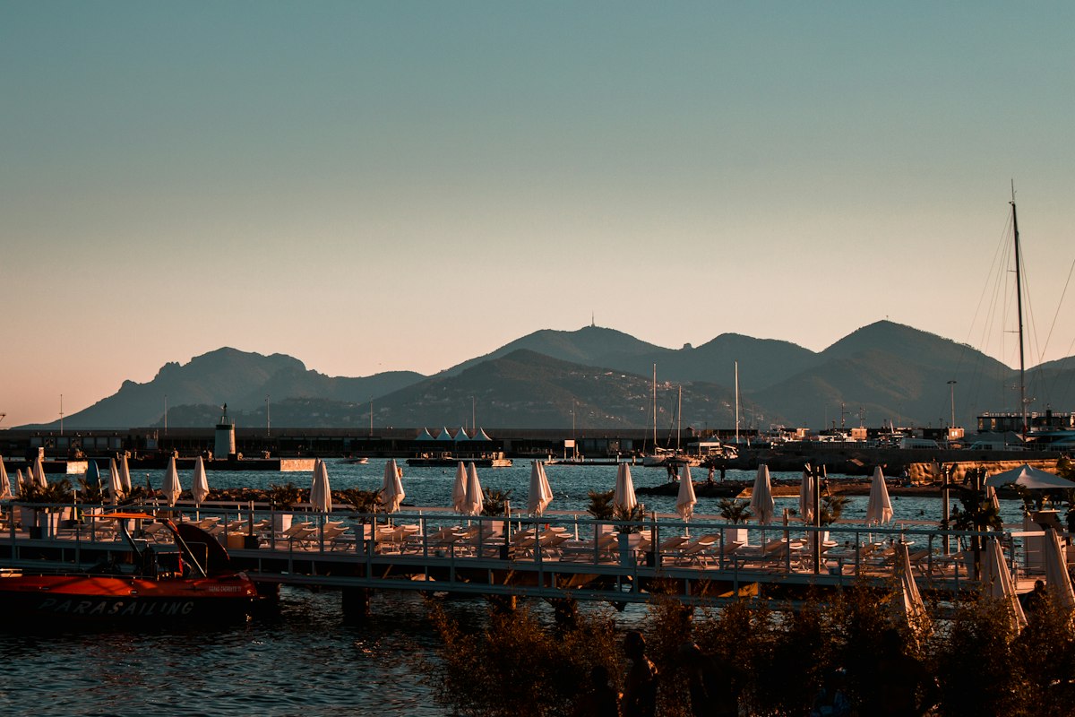The bay of Cannes at golden hour, a private beach platform on piles with rows of folded white parasols and sun loungers, a red parasailing boat moored on the left, a breakwater with a small lighthouse, sailing masts on the right and the Esterel ridge in blue-grey silhouette across the bay