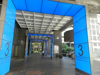 A modern entranceway with bright blue paneling and large numbers '3' displayed on both sides. The design integrates glass and metal with an open hallway extending inward. A person wearing a business outfit is walking toward the building.