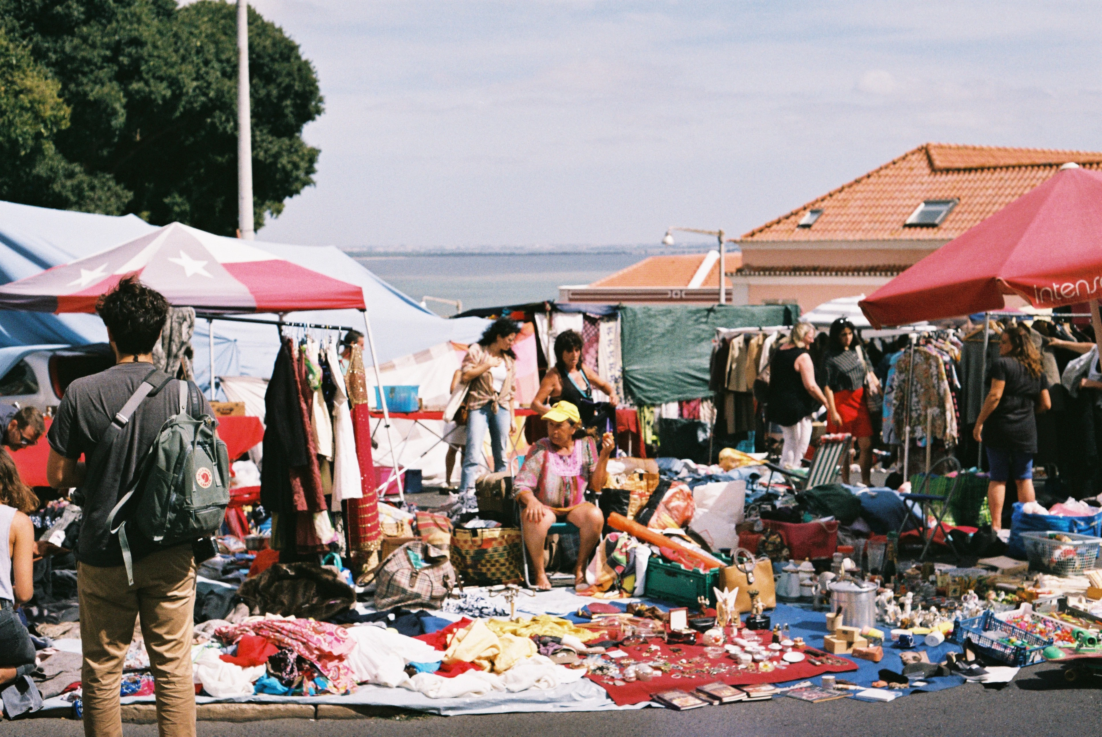 Stalls and blankets of second-hand goods spread across the sunlit ground at the Feira da Ladra flea market in Lisbon, red and white awnings above racks of clothes, browsing shoppers, and the flat blue Tagus estuary behind
