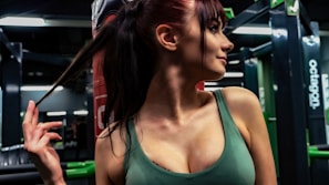 A person with long dark hair is in a gym or fitness center, holding their ponytail. The individual is wearing a sleeveless green top. The background shows gym equipment and mirrors.