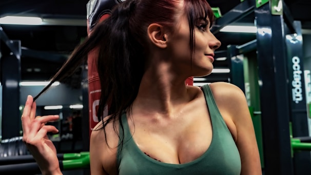 A person with long dark hair is in a gym or fitness center, holding their ponytail. The individual is wearing a sleeveless green top. The background shows gym equipment and mirrors.