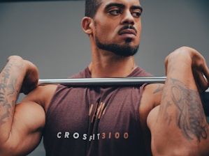 A muscular man with tattoos is holding a barbell across his shoulders. He is wearing a sleeveless shirt with the word 'CROSSFIT3100' printed on it. The background is neutral, focusing attention on the man and his intense expression.