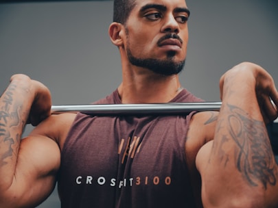 A muscular man with tattoos is holding a barbell across his shoulders. He is wearing a sleeveless shirt with the word 'CROSSFIT3100' printed on it. The background is neutral, focusing attention on the man and his intense expression.