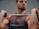 A muscular man with tattoos is holding a barbell across his shoulders. He is wearing a sleeveless shirt with the word 'CROSSFIT3100' printed on it. The background is neutral, focusing attention on the man and his intense expression.