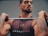 A muscular man with tattoos is holding a barbell across his shoulders. He is wearing a sleeveless shirt with the word 'CROSSFIT3100' printed on it. The background is neutral, focusing attention on the man and his intense expression.