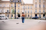 A shirtless man in jeans and green socks stands in a skateboard park, looking away from the camera. A skateboard lies on the ground beside him. In the background, there are classic European-style buildings with rows of windows and balconies, and several parked cars along the street.