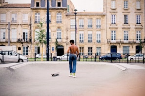 A shirtless man in jeans and green socks stands in a skateboard park, looking away from the camera. A skateboard lies on the ground beside him. In the background, there are classic European-style buildings with rows of windows and balconies, and several parked cars along the street.