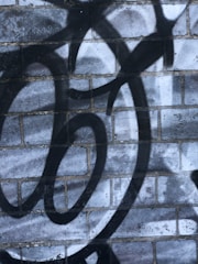 A brick wall features black spray-painted graffiti with circular and swirl patterns. The background bricks are grayish-blue with varying shades and textures.