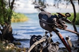 A bicycle equipped with a handlebar bag and other accessories is parked by a lakeside surrounded by trees. The water is calm, and there is vegetation along the shoreline, creating a serene and natural atmosphere.