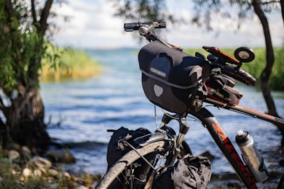 A bicycle equipped with a handlebar bag and other accessories is parked by a lakeside surrounded by trees. The water is calm, and there is vegetation along the shoreline, creating a serene and natural atmosphere.