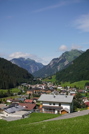 A picturesque mountain village nestled in a lush green valley, surrounded by steep hills and distant peaks. The village features charming houses with colorful roofs, while the foreground is filled with vibrant green grass. The sky is bright and blue with a few scattered clouds.