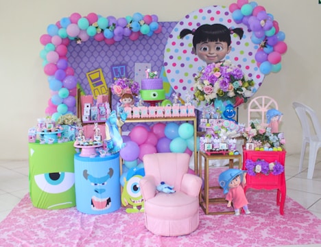The setting displays a colorful children's party with a cartoon theme, featuring balloons in pastel colors like pink, purple, blue, and green. A large poster of a girl with pigtails is part of the backdrop, surrounded by more balloons and flowers. Various themed decorations and toys are arranged on tables, including cartoon characters, gift bags, and treat boxes. The room's bright and cheerful atmosphere is enhanced by a pink armchair and a vibrant pink rug.