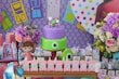 A colorful party setup featuring a two-tiered cake decorated with a large eye and smaller figures on top. The backdrop consists of vibrant geometric patterns and door illustrations. Surrounding the cake are flowers and several themed party items, including gift bags, a plush doll, and themed letters spelling 'Antonella'.