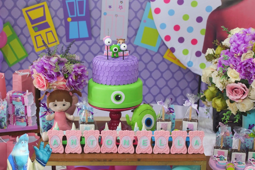 A colorful party setup featuring a two-tiered cake decorated with a large eye and smaller figures on top. The backdrop consists of vibrant geometric patterns and door illustrations. Surrounding the cake are flowers and several themed party items, including gift bags, a plush doll, and themed letters spelling 'Antonella'.