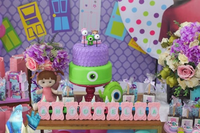 A colorful party setup featuring a two-tiered cake decorated with a large eye and smaller figures on top. The backdrop consists of vibrant geometric patterns and door illustrations. Surrounding the cake are flowers and several themed party items, including gift bags, a plush doll, and themed letters spelling 'Antonella'.