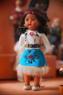 A doll with dark hair and a colorful outfit stands in the foreground. Its attire includes a blue skirt adorned with beaded patterns, a white top with fluffy cuffs, and a red headband, all embellished with multicolored beads. The background is blurred, featuring other dolls or figures.