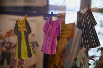 Several paper cutouts of robes are clipped to a string with colorful clothespins. Each robe features distinct designs, with one in black and yellow, another in purple, and the last one in black and white stripes. The background contains blurred images and colors, suggesting a display of illustrations or costumes.