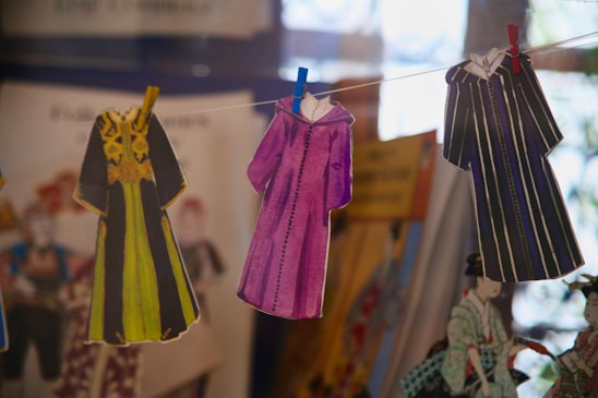 Several paper cutouts of robes are clipped to a string with colorful clothespins. Each robe features distinct designs, with one in black and yellow, another in purple, and the last one in black and white stripes. The background contains blurred images and colors, suggesting a display of illustrations or costumes.