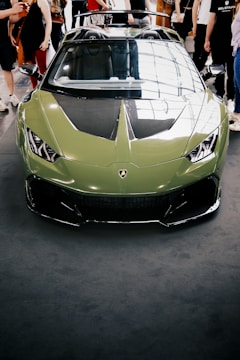A green sports car with a sleek, aerodynamic design is parked indoors. The car has a shiny exterior with a prominent front grille and sharp headlights. Multiple people are standing around the car, some with their hands in their pockets and others holding bags. The image reflects light from large windows, creating interesting reflections on the car's surface.