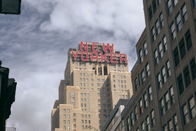 A tall building prominently displays the words 'NEW YORKER' in red letters against a mostly overcast sky. The architecture features a series of stepped, art-deco style tiers and is flanked by other high-rise buildings. The perspective offers a view upwards, emphasizing the height and grandeur of the structure.