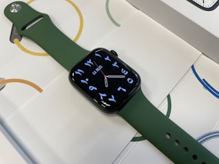 A smart watch with a sleek, dark green strap and a digital display featuring Arabic numerals and a minimalistic watch face. The watch is placed on a surface with a pattern of circles and rope-like designs in various colors.