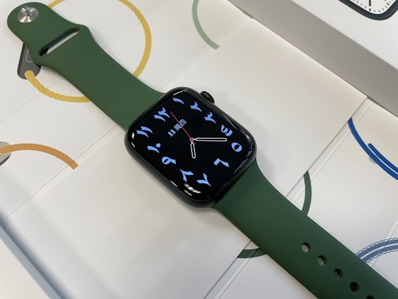 A smart watch with a sleek, dark green strap and a digital display featuring Arabic numerals and a minimalistic watch face. The watch is placed on a surface with a pattern of circles and rope-like designs in various colors.