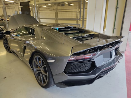 A sleek, high-performance sports car with a futuristic design, parked indoors. The car has an angular, aerodynamic body, large air intakes, and a prominent rear wing. The rear engine cover is open, and the car's glossy finish reflects the lights from the surroundings.