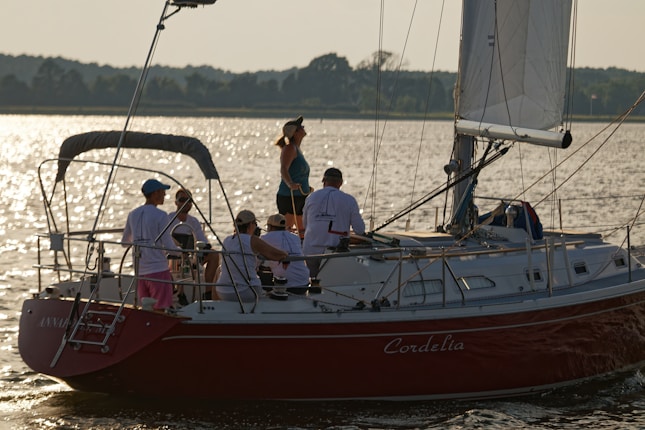 Several people are gathered on a sailboat named 'Cordelia' as it sails on a calm body of water. The sun is setting, casting a warm glow over the scene. The group appears relaxed and is dressed in casual summer clothing.