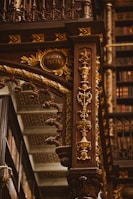 An intricately designed wooden column adorned with ornate carvings in gold, featuring floral patterns and elegant details. The background includes shelves filled with books, highlighting the setting in an opulent library. The column includes an inscription reading 'Rocha Pitta'.
