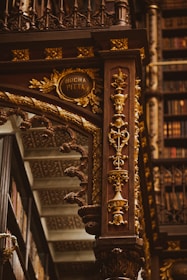An intricately designed wooden column adorned with ornate carvings in gold, featuring floral patterns and elegant details. The background includes shelves filled with books, highlighting the setting in an opulent library. The column includes an inscription reading 'Rocha Pitta'.