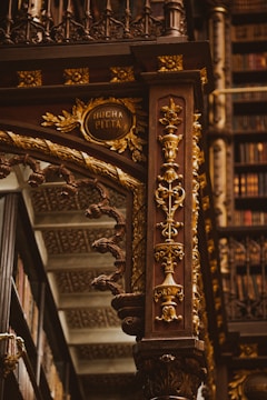 An intricately designed wooden column adorned with ornate carvings in gold, featuring floral patterns and elegant details. The background includes shelves filled with books, highlighting the setting in an opulent library. The column includes an inscription reading 'Rocha Pitta'.