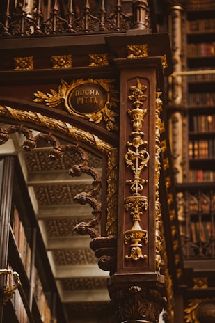 An intricately designed wooden column adorned with ornate carvings in gold, featuring floral patterns and elegant details. The background includes shelves filled with books, highlighting the setting in an opulent library. The column includes an inscription reading 'Rocha Pitta'.