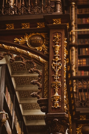 An intricately designed wooden column adorned with ornate carvings in gold, featuring floral patterns and elegant details. The background includes shelves filled with books, highlighting the setting in an opulent library. The column includes an inscription reading 'Rocha Pitta'.