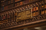 A library wall filled with books on shelves, with a detailed and ornate metal railing in front. Mounted on the rail is a plaque dedicated to Machado de Assis, a Brazilian writer, commemorating his memory.