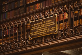 A library wall filled with books on shelves, with a detailed and ornate metal railing in front. Mounted on the rail is a plaque dedicated to Machado de Assis, a Brazilian writer, commemorating his memory.